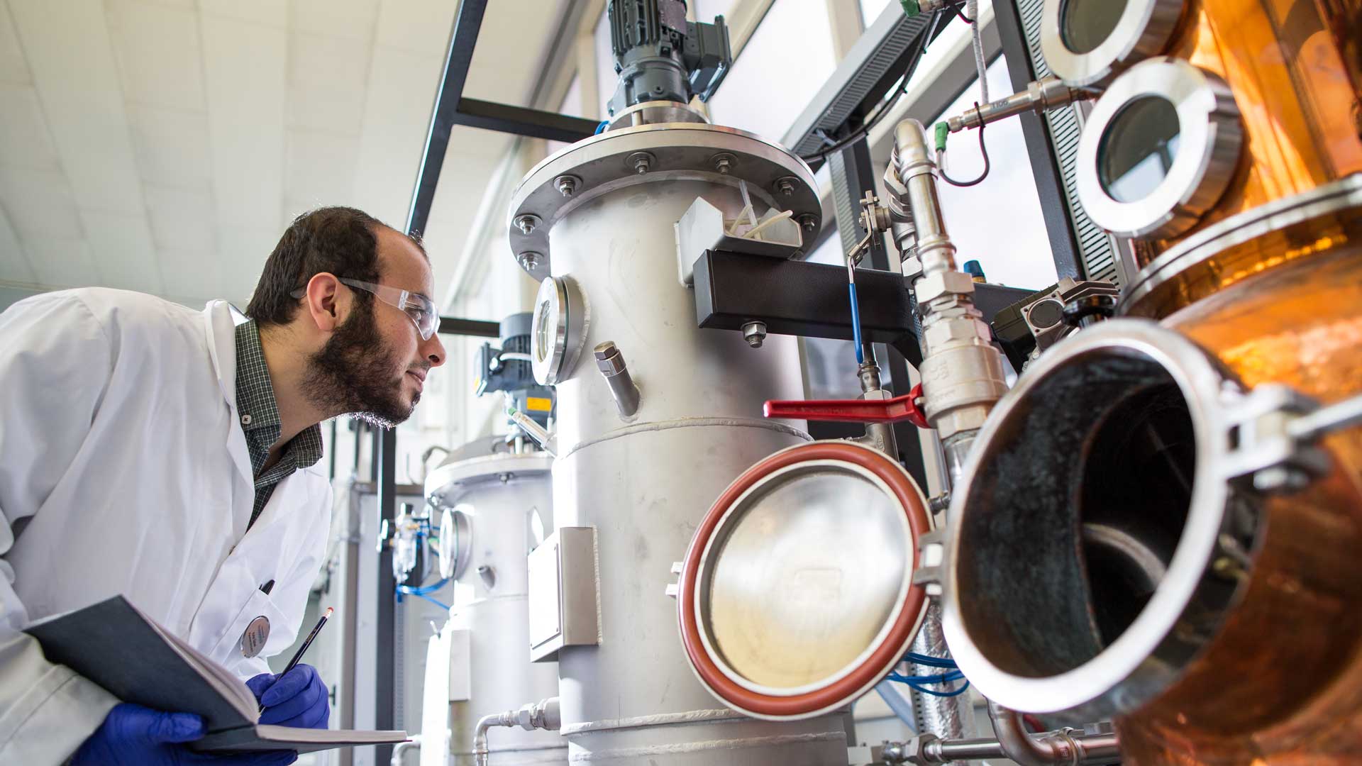 Someone in a lab coat looking at a piece of equipment and taking notes.