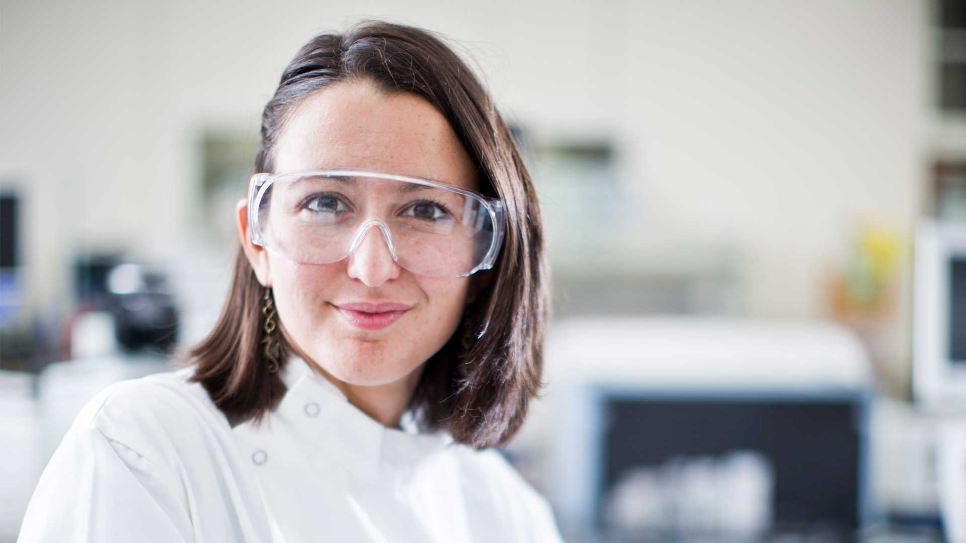 A scientist sits and looks directly at the camera. She is wearing a lab coat and protective goggles. There is a lab in the background.
